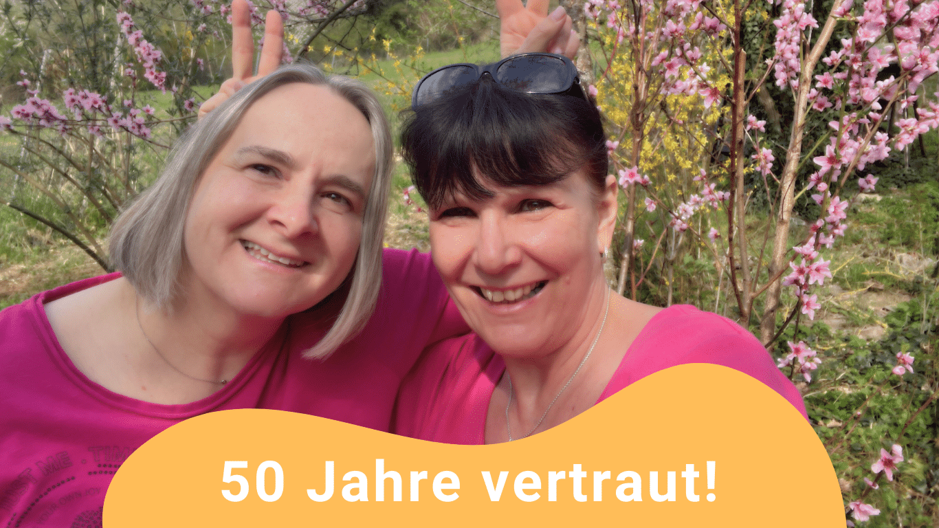 50 Jahre Freundschaft verbindet Sandra und ihre Freundin Britta. Sie sind zusammen auf dem Foto und haben sich gegenseitig umarmt und blicken lachend in die Kamera. Sie stehen vor blühenden Weinbergspfirsichbäumen und haben sich gegenseitig mit den Händen hinter dem Kopf Hasenohren gemacht.