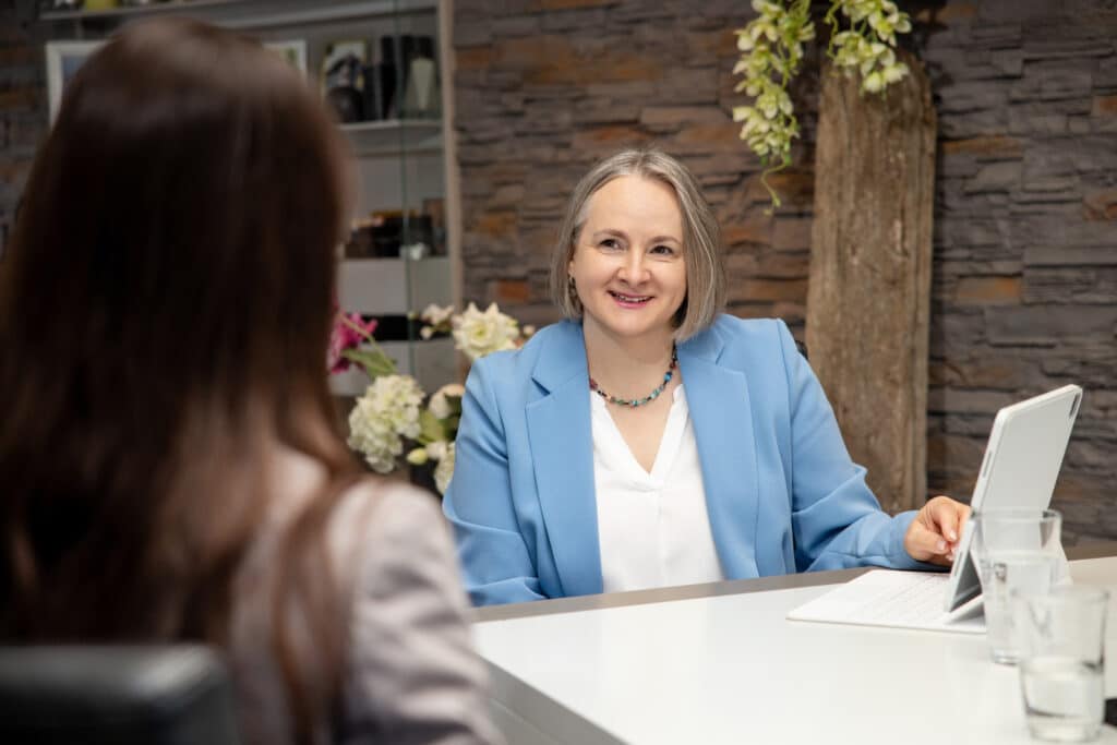 Alexandra Kärgel in hellblauem Blazer mit weißer Bluse hört einer Unternehmerin in stilvoller Beratungsatmosphäre mit Steinwand, Holzdeko und Blumen aufmerksam zu – Beratungsgespräch zur Unternehmensnachfolge in Cochem an der Mosel