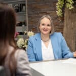 Alexandra Kärgel in hellblauem Blazer mit weißer Bluse hört einer Unternehmerin in stilvoller Beratungsatmosphäre mit Steinwand, Holzdeko und Blumen aufmerksam zu – Beratungsgespräch zur Unternehmensnachfolge in Cochem an der Mosel