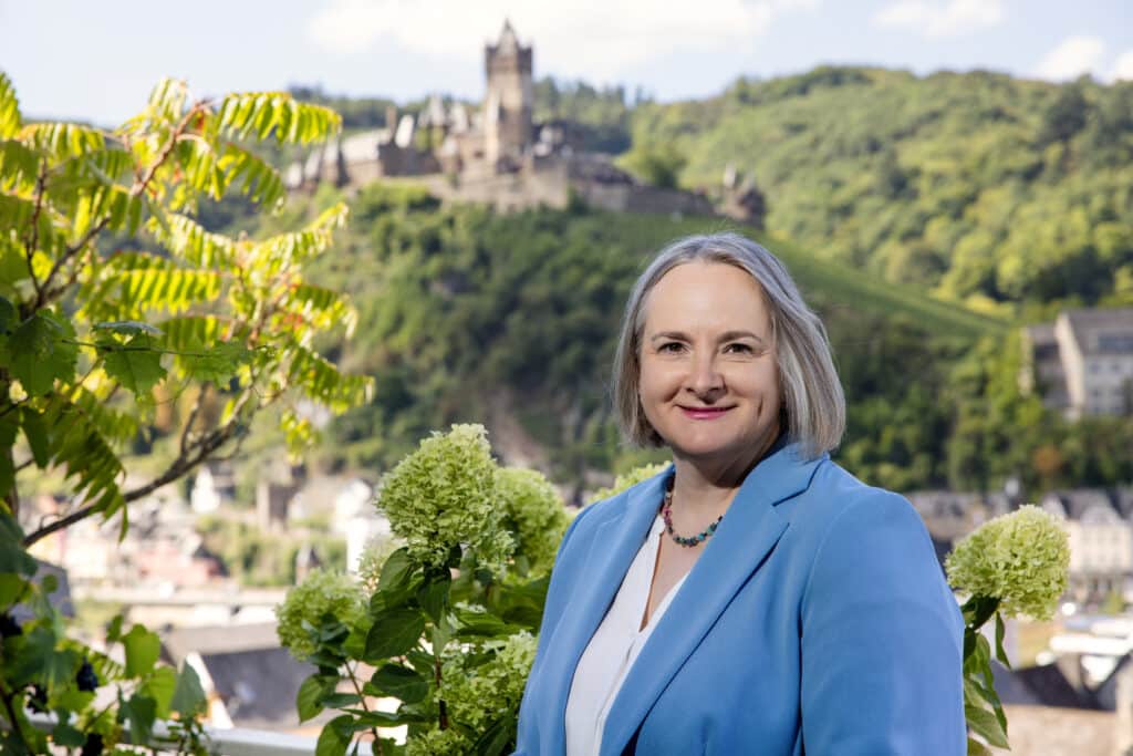 Alexandra Kärgel, Expertin für Unternehmensnachfolge, lächelnd in hellblauem Blazer an einem Sommertag vor der Burg Cochem an der Mosel