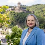 Alexandra Kärgel, Expertin für Unternehmensnachfolge, lächelnd in hellblauem Blazer an einem Sommertag vor der Burg Cochem an der Mosel