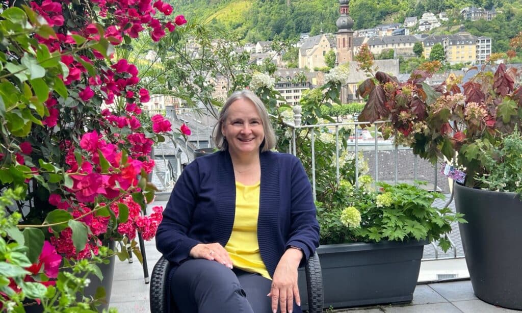 Alexandra Kärgel auf ihrem blühenden Balkon. Sie trägt ein gelbes Shirt mit einer blauen Shirtjacke