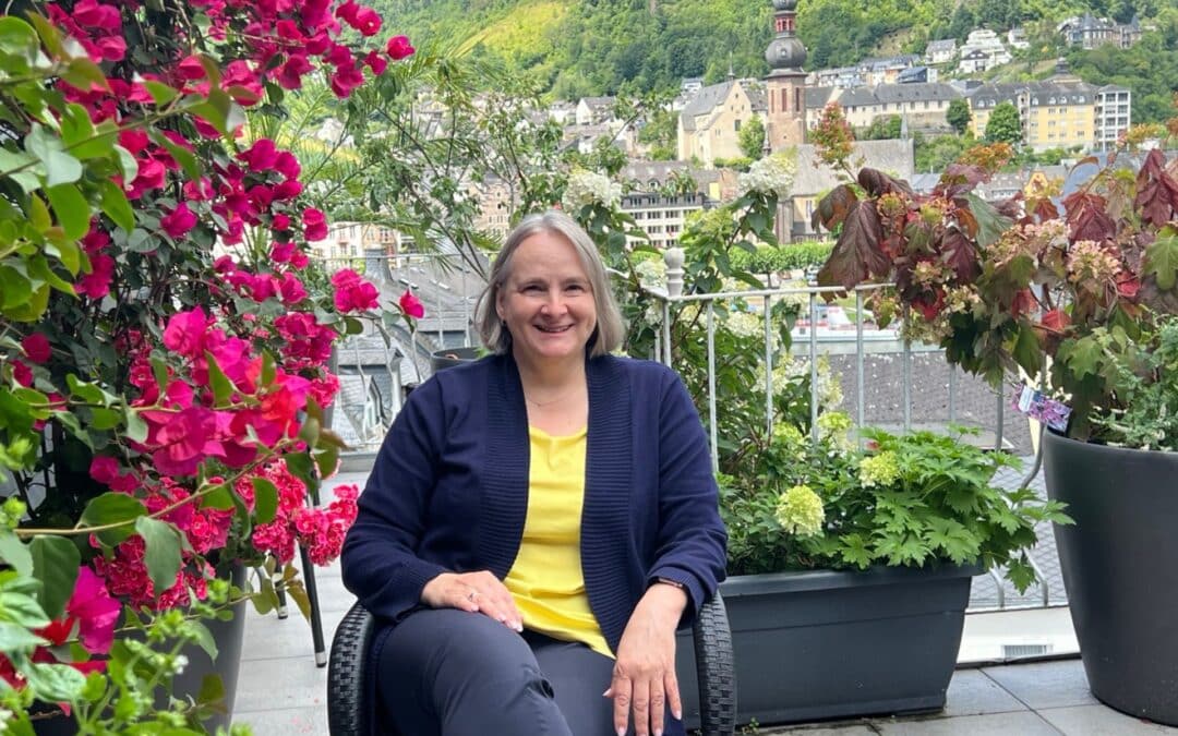 Alexandra Kärgel auf ihrem blühenden Balkon. Sie trägt ein gelbes Shirt mit einer blauen Shirtjacke
