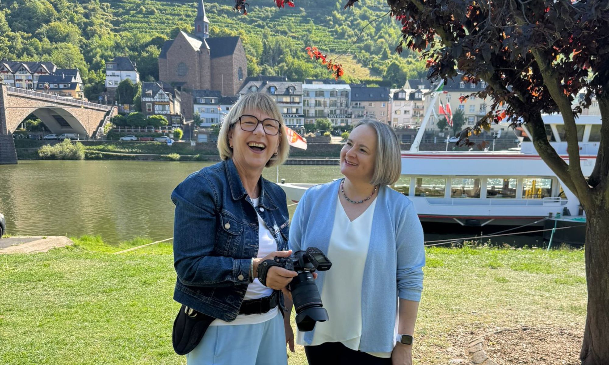 Fotografin Karina Schuh und Alexandra Kärgel zusammen beim Fotoshooting an der Mosel. Sie sind in blauen Tönen gekleidet und lachen zusammen. Sie stehen auf der Wiese am Moselufer.