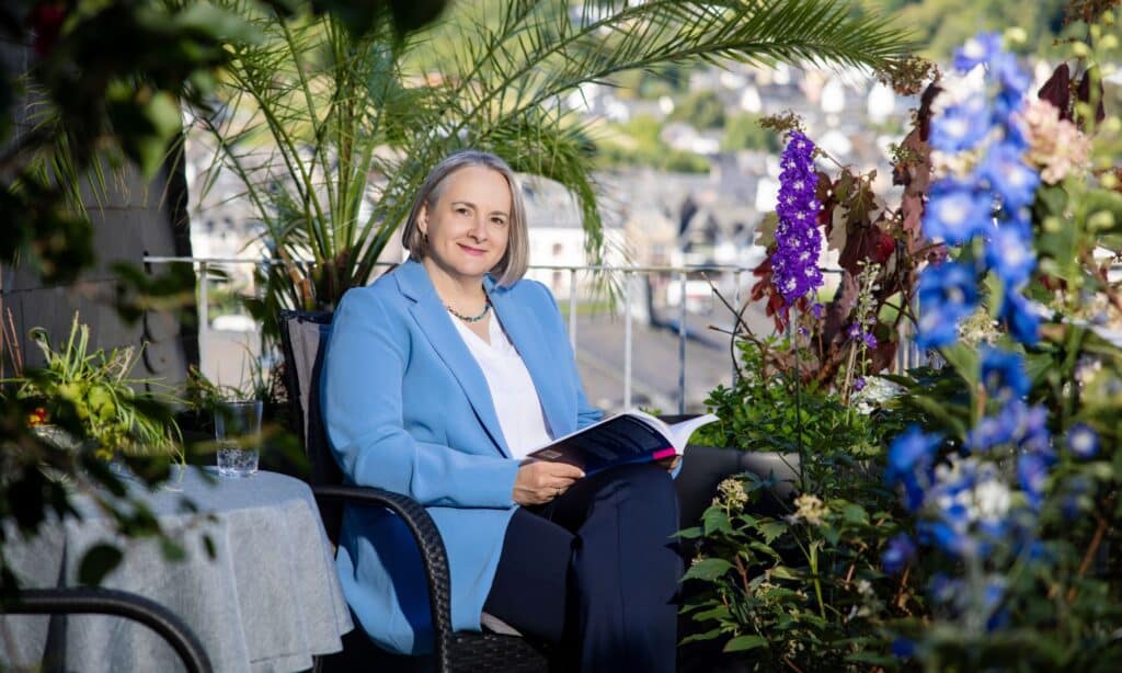 Alexandra Kärgel sitzt auf ihrem Balkon umgeben von blühenden Pflanzen. Sie trägt einen hellblauen Blazer, eine weiße Bluse und eine dunkelblaue Hose. Auf dem Schoß hat sie ein Hose.