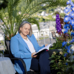 Alexandra Kärgel liest ihr Buch auf einer Terrasse – Nachfolgeberaterin und Autorin. Um sie herum sind viele bunte Pflanzen. Sie trägt einen hellblauen Blazer und eine dunkelblaue Hose.
