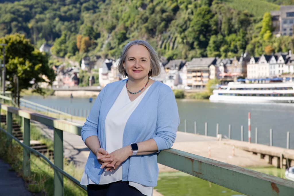 Alexandra Kärgel, Beraterin für Unternehmensnachfolge, lächelt an einem Geländer am Flussufer vor der Kulisse der malerischen Kleinstadt Cochem an der Mosel. Sie trägt eine hellblaue Strickweste und eine weiße Bluse.