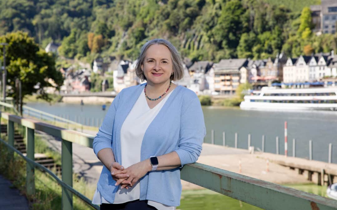 Alexandra Kärgel, Beraterin für Unternehmensnachfolge, lächelt an einem Geländer am Flussufer vor der Kulisse der malerischen Kleinstadt Cochem an der Mosel. Sie trägt eine hellblaue Strickweste und eine weiße Bluse.
