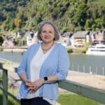 Alexandra Kärgel, Beraterin für Unternehmensnachfolge, lächelt an einem Geländer am Flussufer vor der Kulisse der malerischen Kleinstadt Cochem an der Mosel. Sie trägt eine hellblaue Strickweste und eine weiße Bluse.