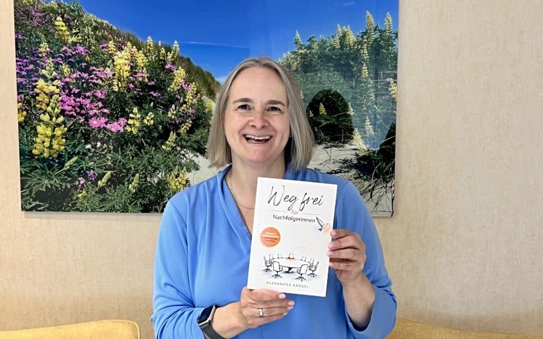 Alexandra Kärgel hält lächelnd ihr Buch „Weg frei für Nachfolgerinnen“, ein Mutmachbuch für Nachfolgerinnen in Familienunternehmen. Sie trägt ein hellblaues Shirt. Im Hintergrund ein Bild an der Wand mit einem blauen Himmel und bunten Blumen.