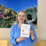 Alexandra Kärgel hält lächelnd ihr Buch „Weg frei für Nachfolgerinnen“, ein Mutmachbuch für Nachfolgerinnen in Familienunternehmen. Sie trägt ein hellblaues Shirt. Im Hintergrund ein Bild an der Wand mit einem blauen Himmel und bunten Blumen.